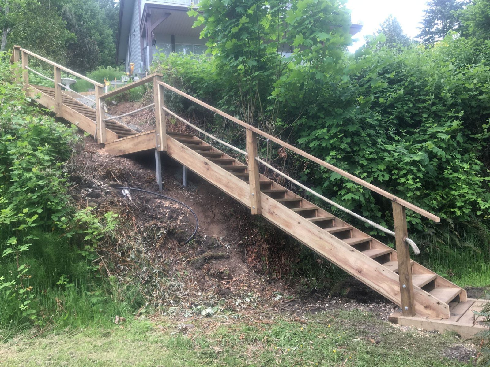 Finished wooden staircase with horizontal cable-style railing and angled landing below a hillside house