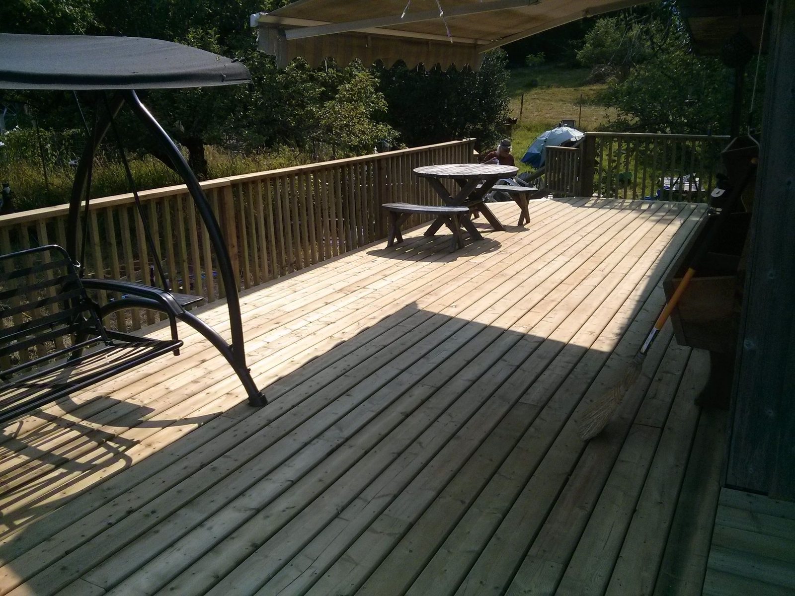 Large sunny backyard wood deck with railing, porch swing and picnic table by a home