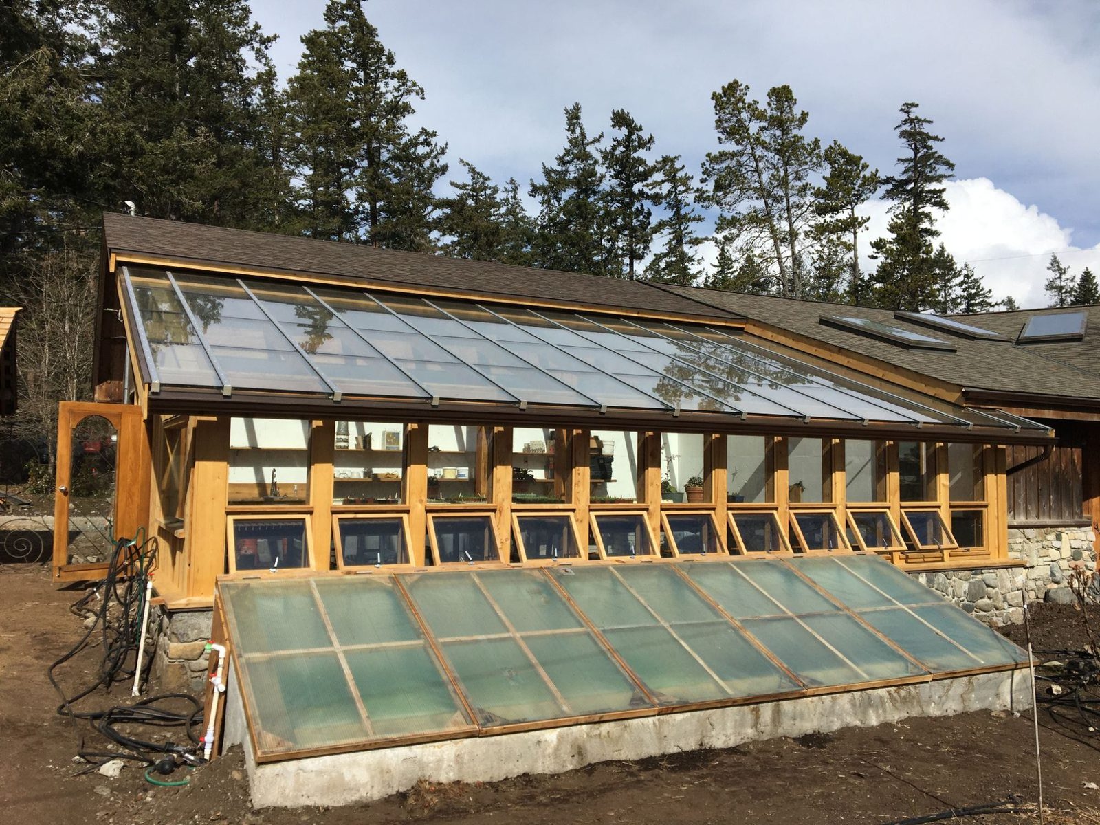 Side view of glass-roofed timber-frame greenhouse with awning windows and angled cold frame on a forested lot, Quadra Island