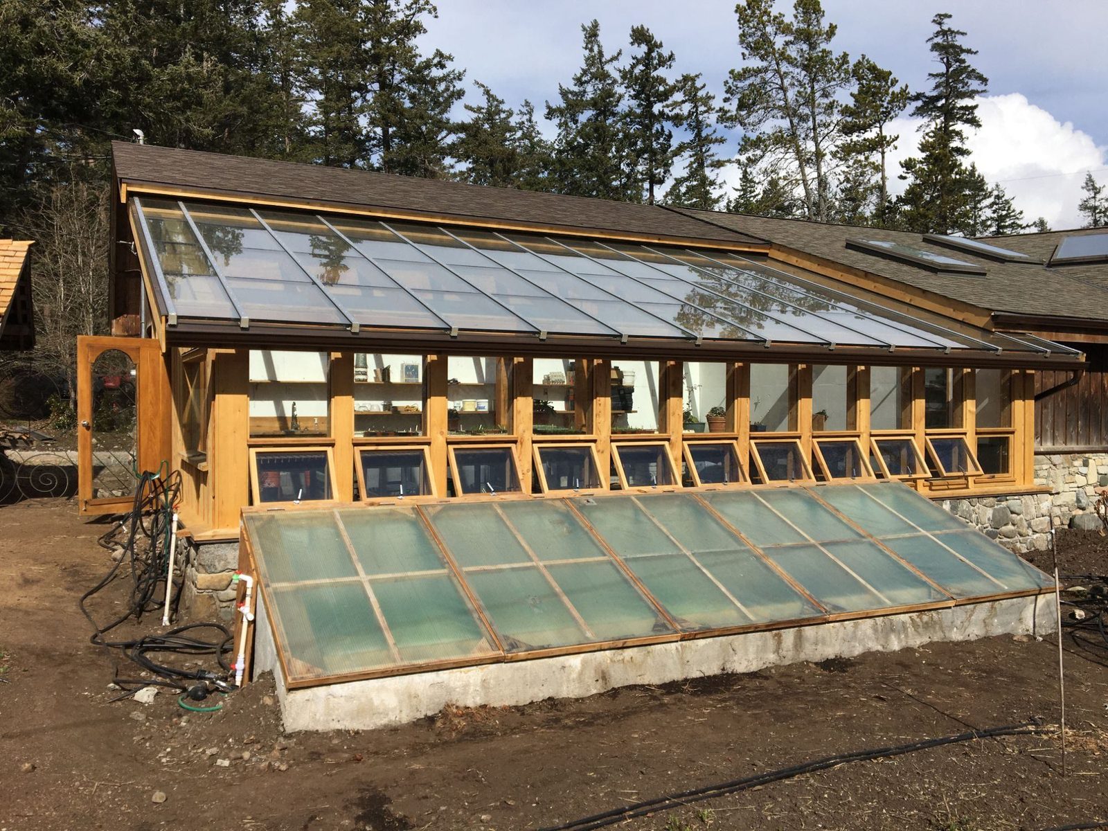 Exterior of timber-frame greenhouse with sloped glass roof, vent windows and glazed cold frame on stone foundation, Quadra Island
