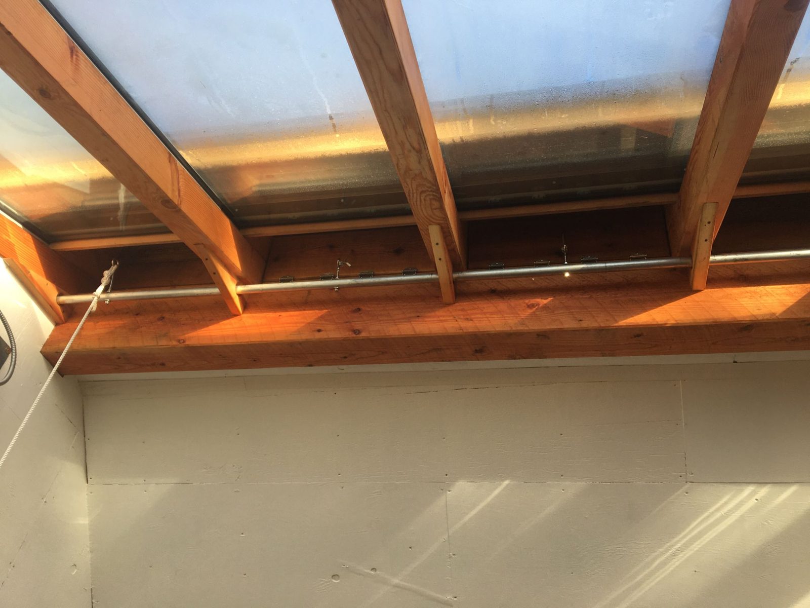 Underside of greenhouse glass roof with sunlit fir rafters and steel tie rod above a plywood wall, Quadra Island