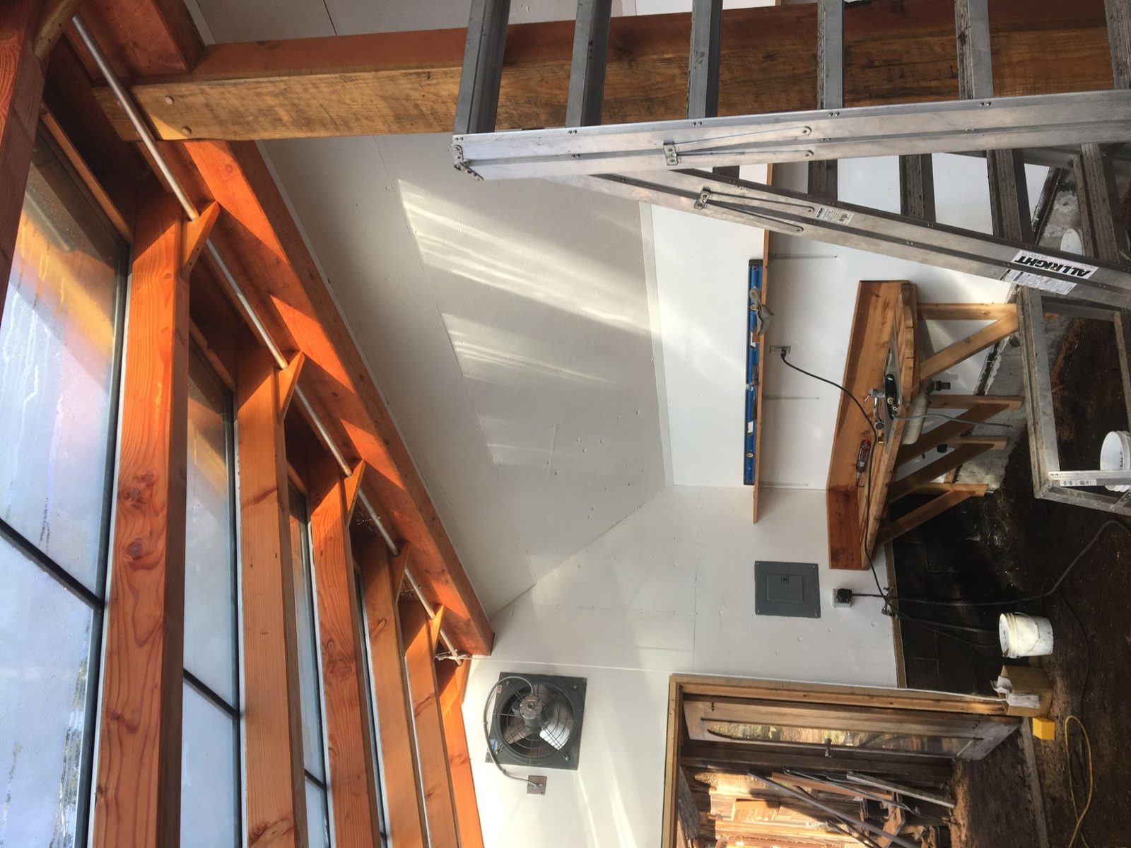 Greenhouse interior during construction with fir posts, exhaust fan, ladder and workbench against white walls, Quadra Island