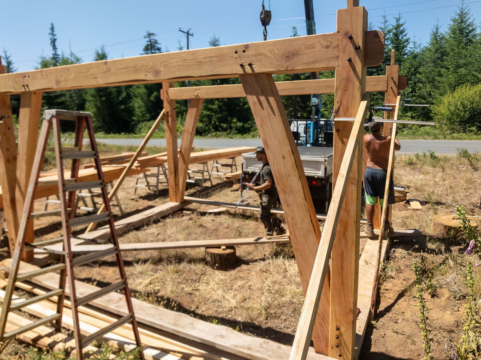 Erected timber frame bent with tie beam and angled knee braces beside ladders on Quadra Island