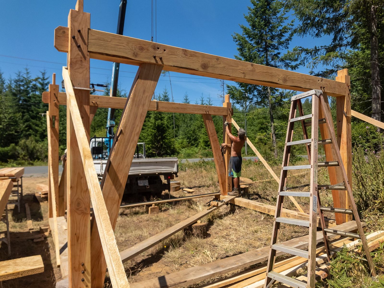 Carpenter aligning a knee brace on a tall timber frame bent with tie beam under a crane on Quadra Island