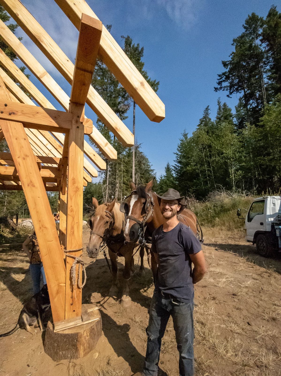 Smiling builder standing with draft horses beside the completed timber frame pavilion on Quadra Island