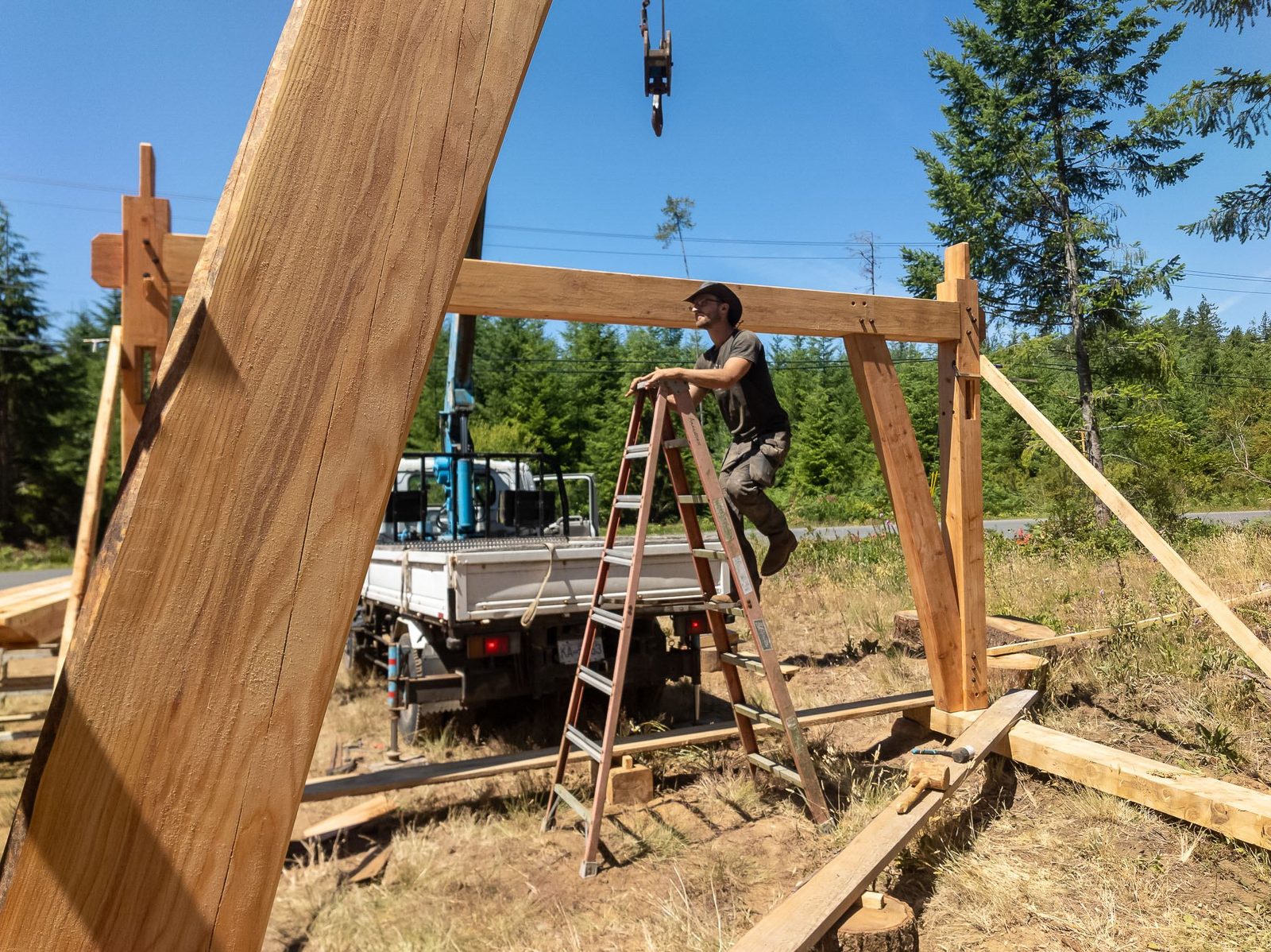 Carpenter on a stepladder working at a raised timber frame bent with crane truck behind on Quadra Island