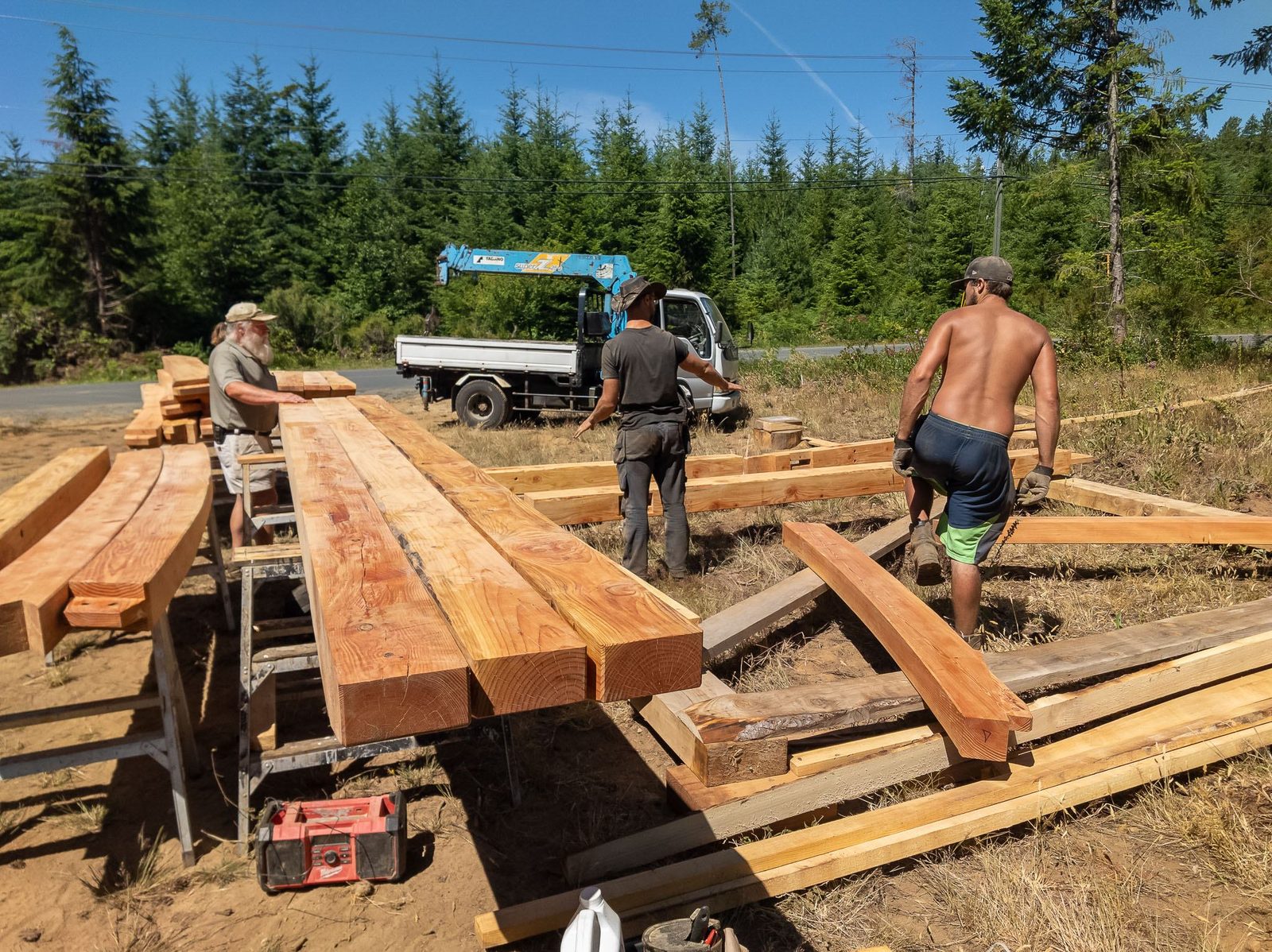 Three carpenters laying out heavy timber frame beams with a crane truck in the clearing on Quadra Island