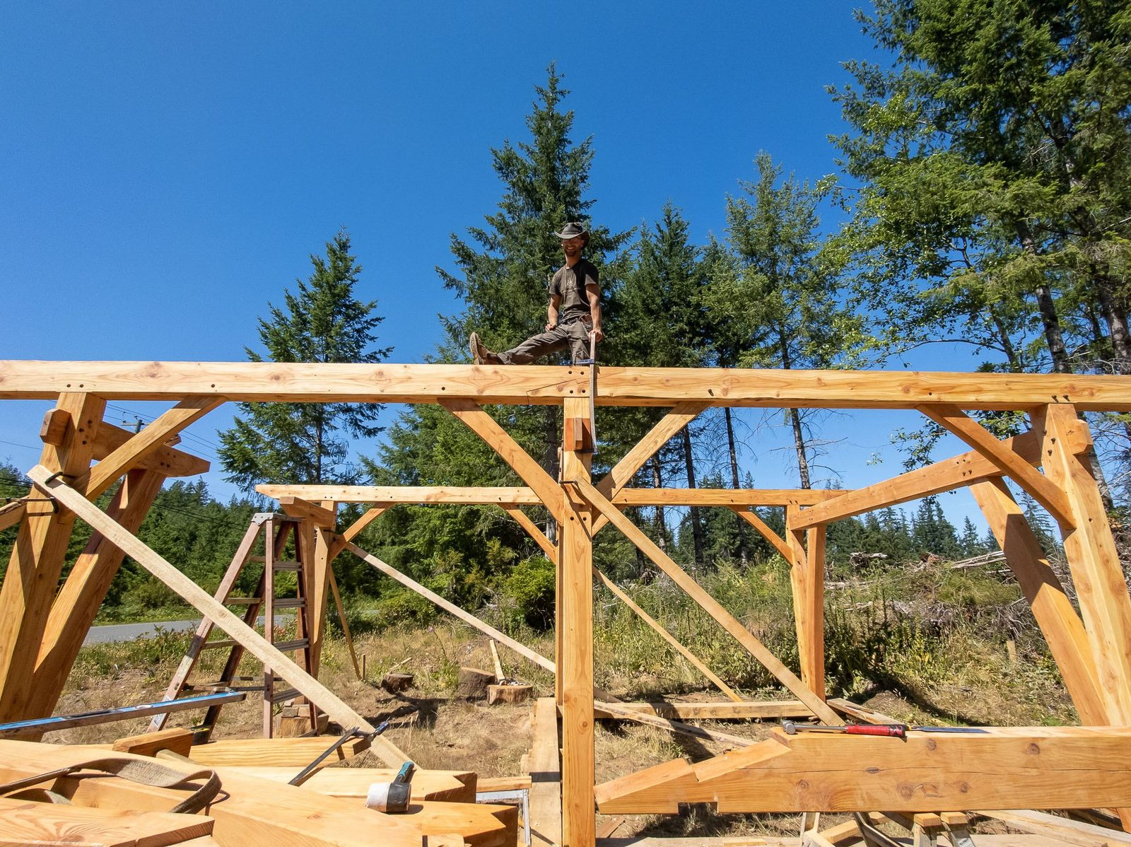 Carpenter sitting on the ridge of a completed timber frame skeleton against blue sky on Quadra Island