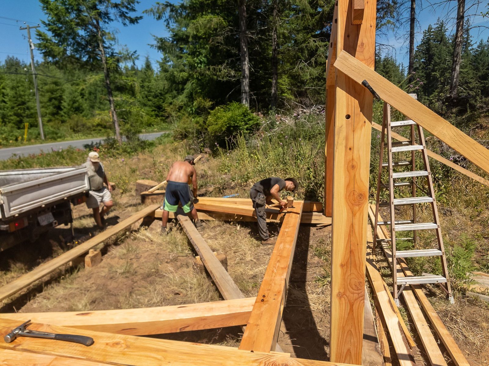 Carpenters fitting timber frame floor members on the ground beside a standing braced post on Quadra Island