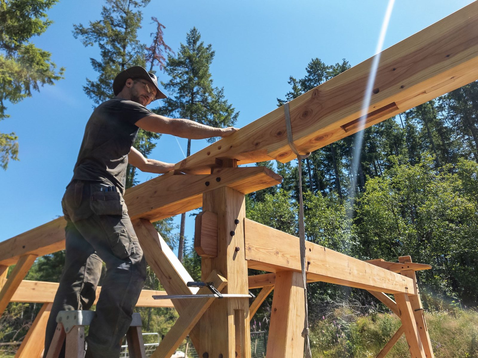 Carpenter on a ladder fitting a rafter along the timber frame purlin against the sky on Quadra Island