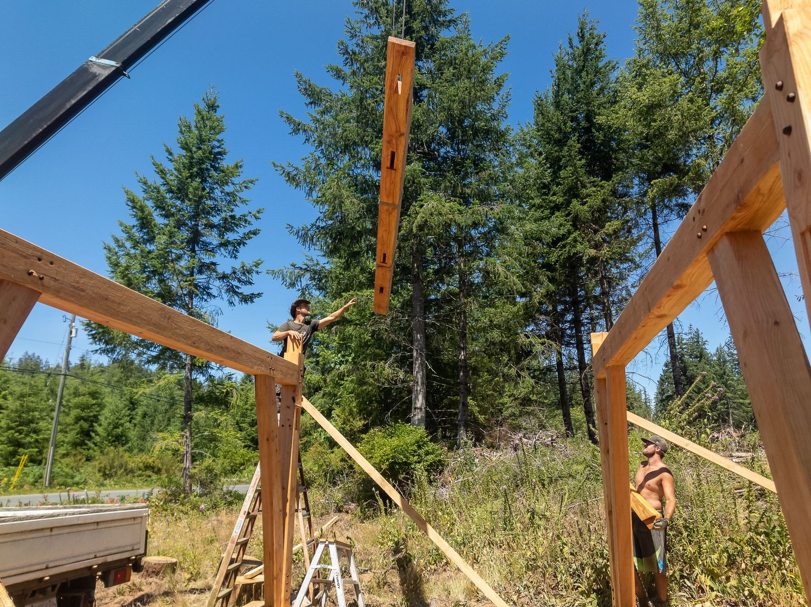 Carpenter on a ladder guiding a tall timber post lifted by crane between two bents on Quadra Island