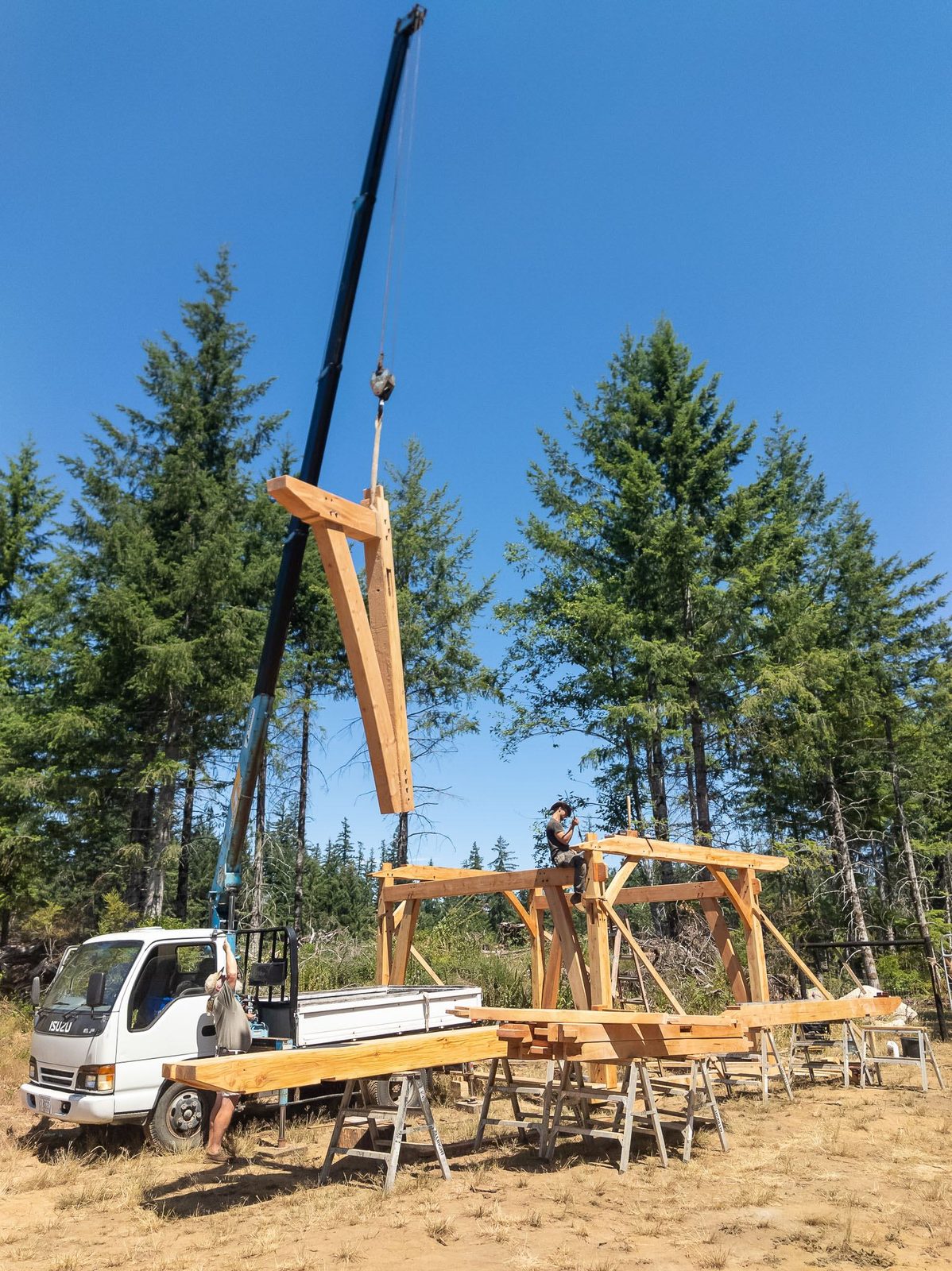 Tall crane hoisting a timber rafter assembly toward the frame with a truck below on Quadra Island