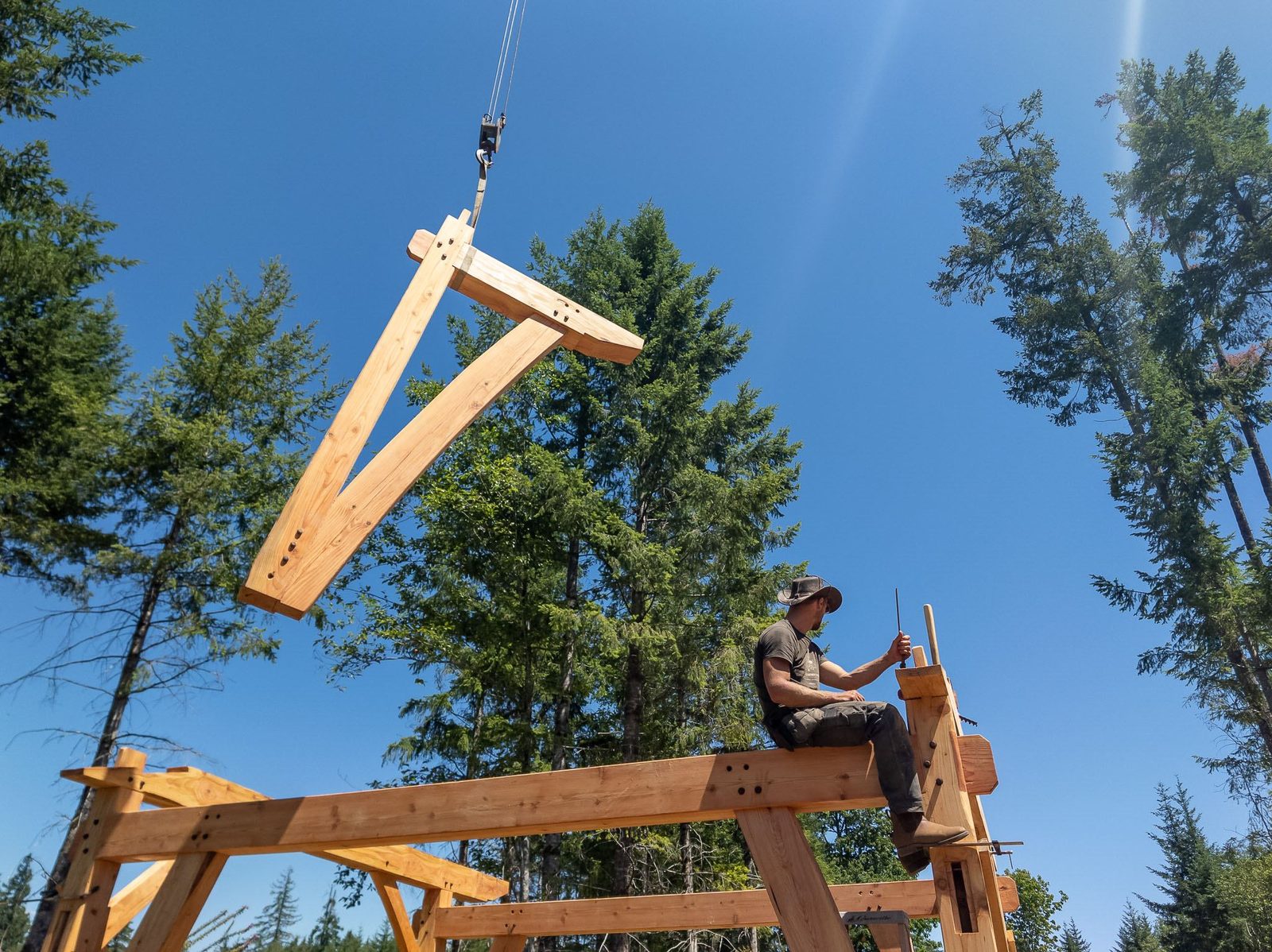 Low-angle view of a crane-lifted timber rafter above a carpenter seated on the frame on Quadra Island