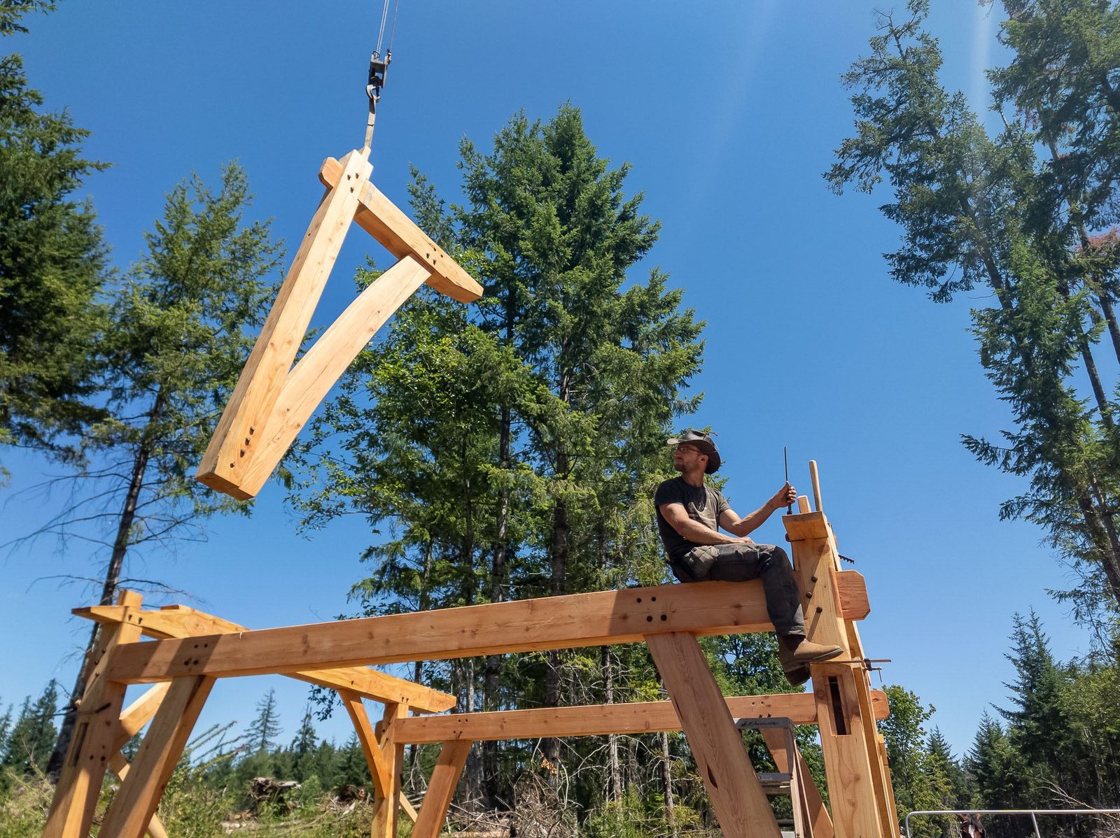 Crane-suspended timber rafter angled over a carpenter on the timber frame against blue sky on Quadra Island