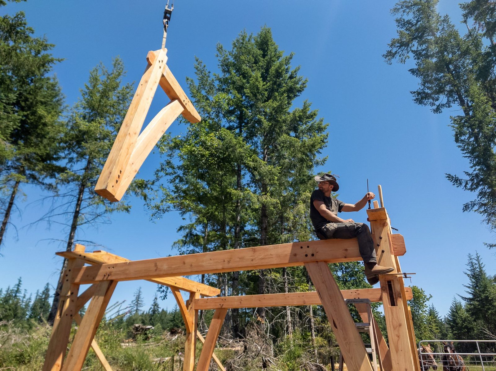 Carpenter seated on the timber frame guiding a crane-lifted rafter overhead on Quadra Island