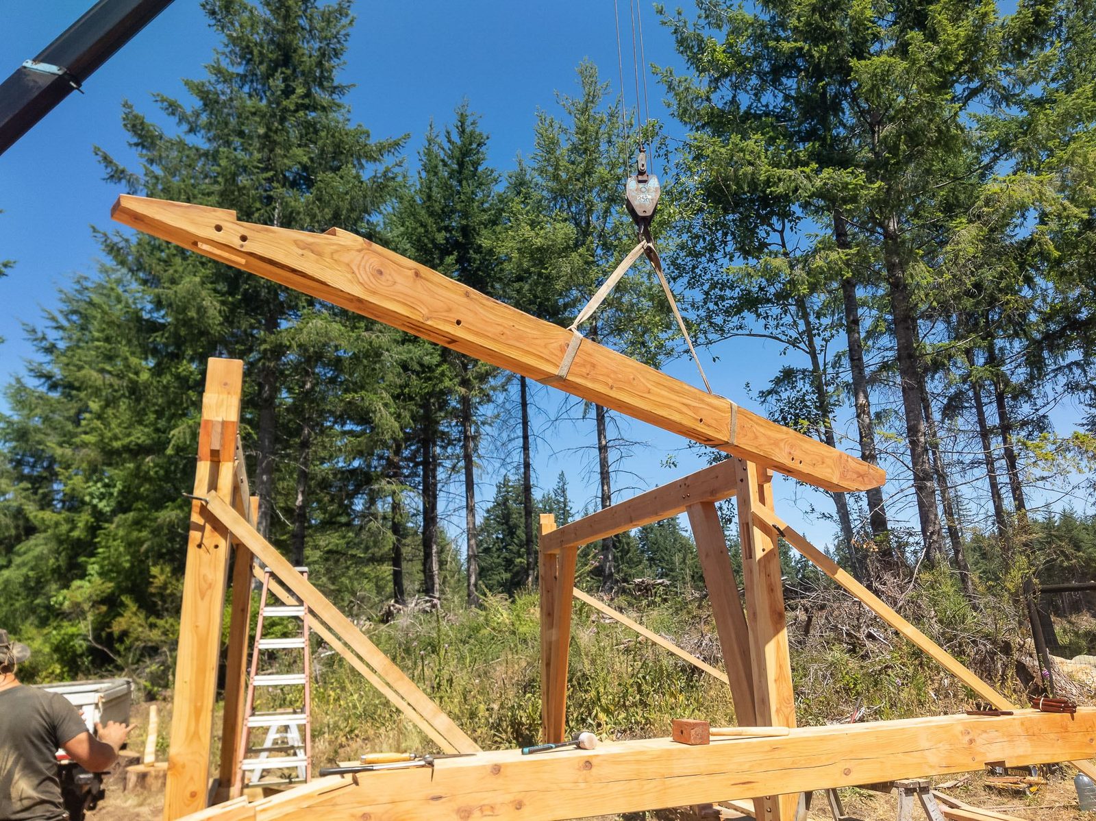 Long timber rafter lifted by crane slings above a partly raised frame against blue sky on Quadra Island