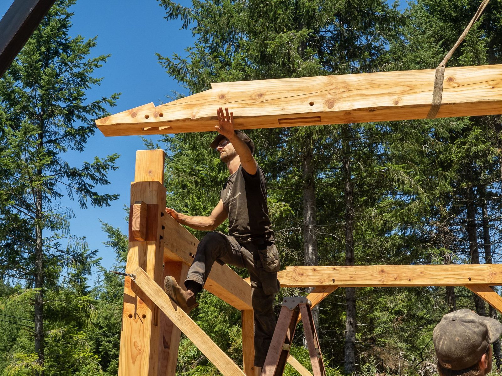Carpenter seated on a frame guiding a crane-lifted rafter onto the top of a timber frame bent on Quadra Island