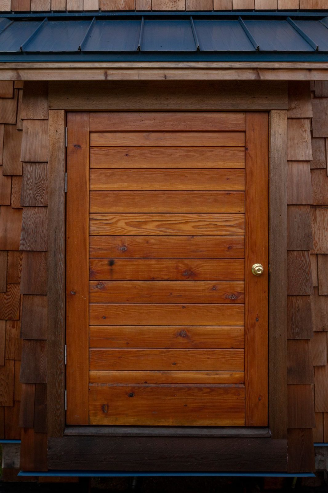 Wood plank entry door framed by cedar shingle siding under a metal roof on a tiny home