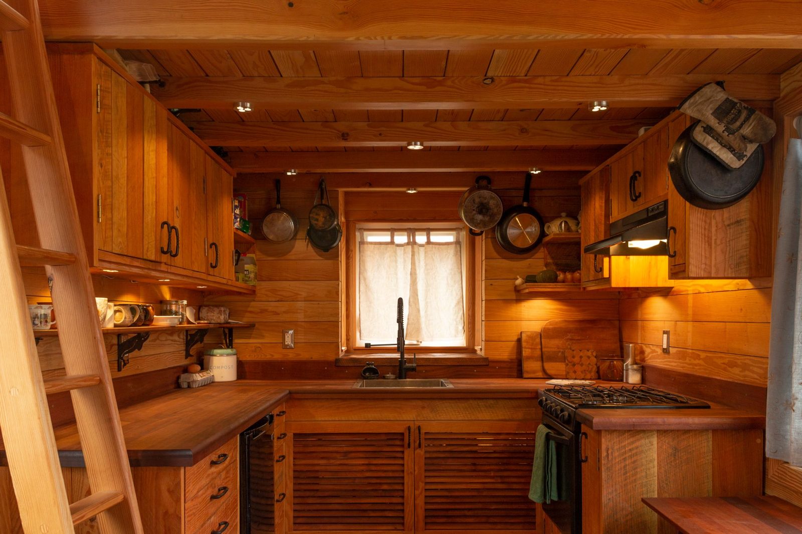 Compact wood kitchen with timber counters, gas range and hanging pans inside a tiny home on Quadra Island