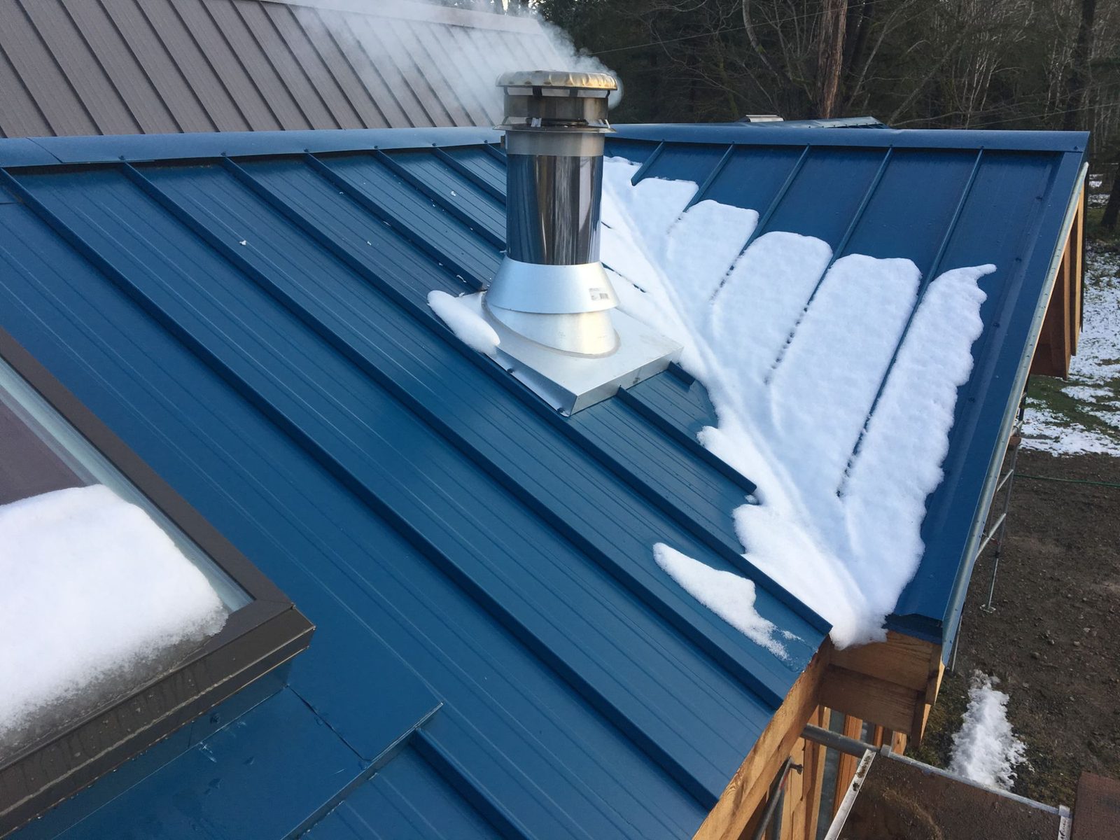 Blue standing-seam metal roof with chimney flue and snow on a tiny home on Quadra Island