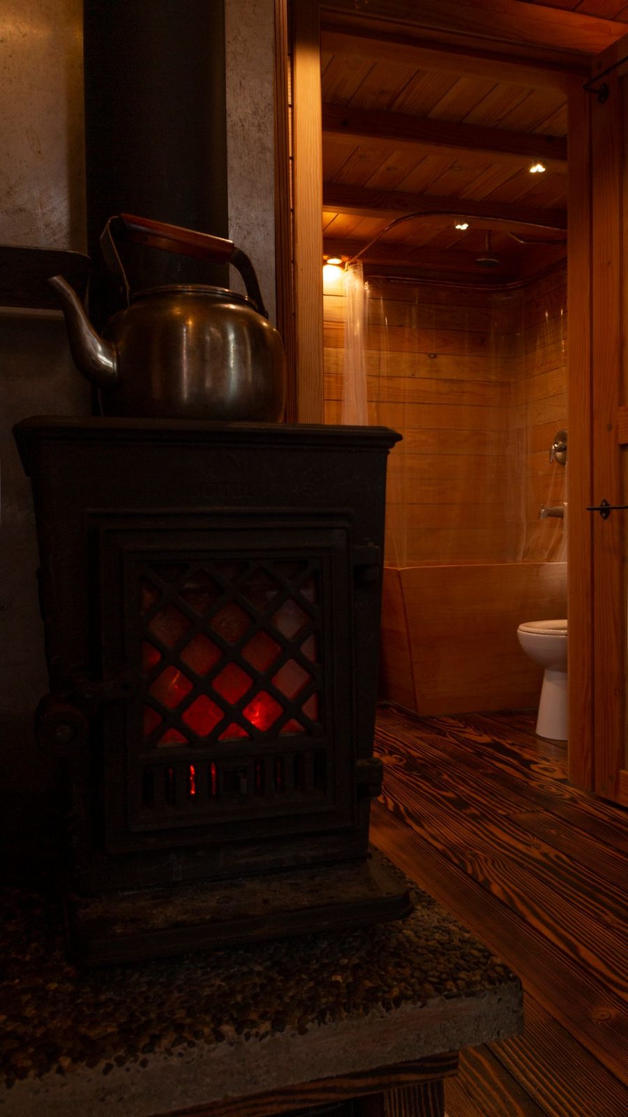 Cast iron wood stove with copper kettle and adjacent wood-clad bathroom in a tiny home